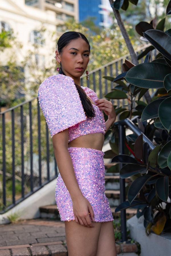 Woman wearing a pink sequin Filipiniana terno standing outdoors with plants and buildings in the background