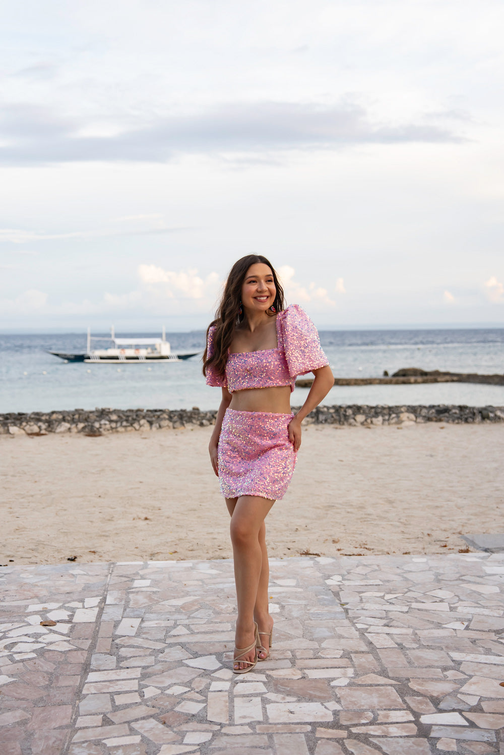 Woman in a pink sequin Filipiniana terno standing on a beach with ocean and sky in the background
