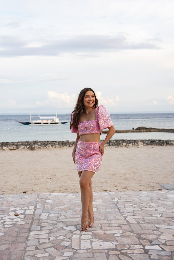 Woman in a pink sequin Filipiniana terno standing on a beach with ocean and sky in the background