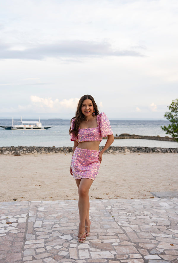 Woman in a pink sequin Filipiniana terno standing on a stone pathway by the beach