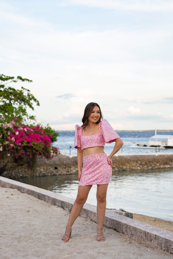 Woman in a pink sequin Filipiniana terno standing by a waterfront with flowers and a boat in the background.
