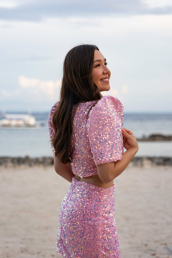 Woman in a pink sequin Filipiniana terno standing on a beach with ocean and sky in the background