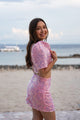 Woman in a pink sequin Filipiniana terno standing on a beach with water and sky in the background