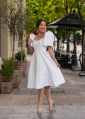 Woman in a white jacquard dress with filipiniana sleeves standing on a sidewalk with trees and outdoor seating in the background.