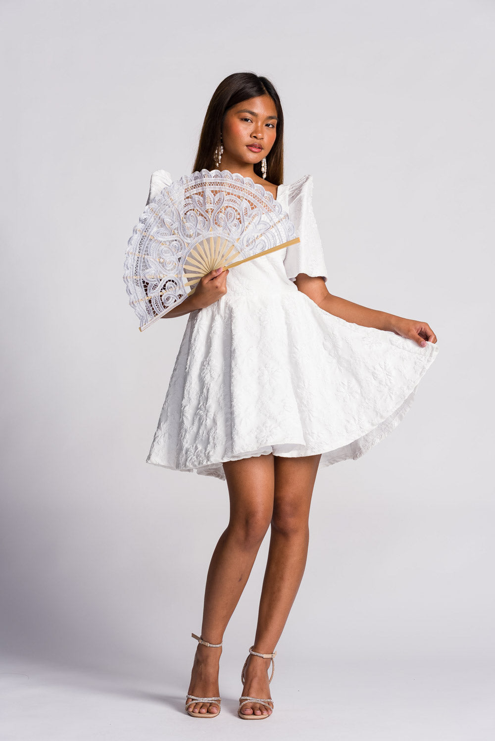 Woman wearing a white filipiniana mini dress holding a lace pay pay fan on a plain background