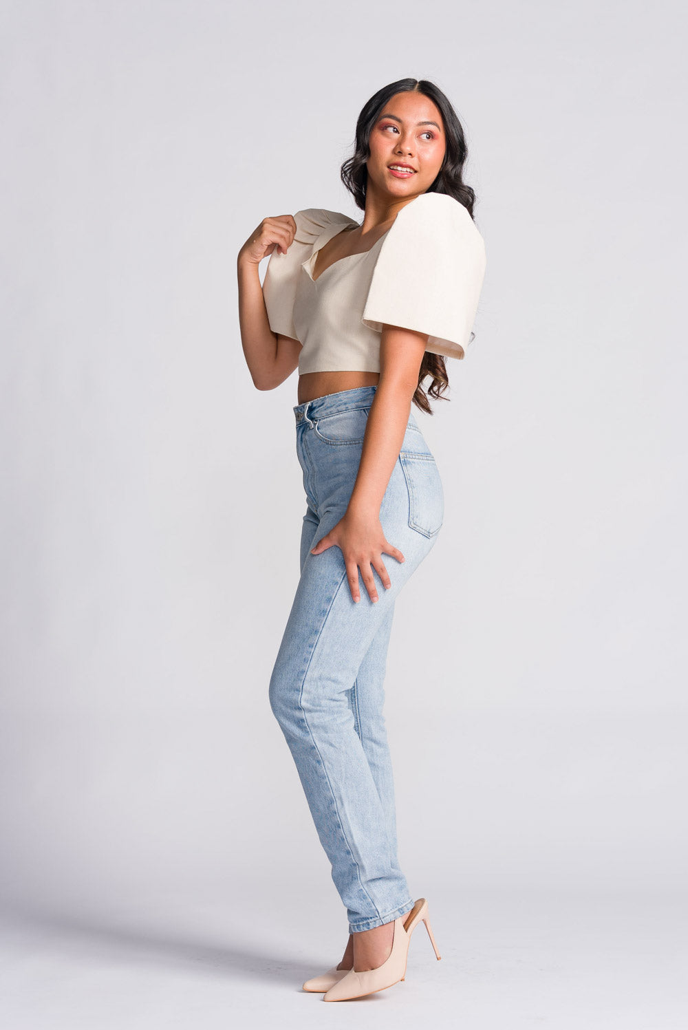 Woman wearing a white cream filipiniana crop top with butterfly sleeves and light blue jeans on a plain background