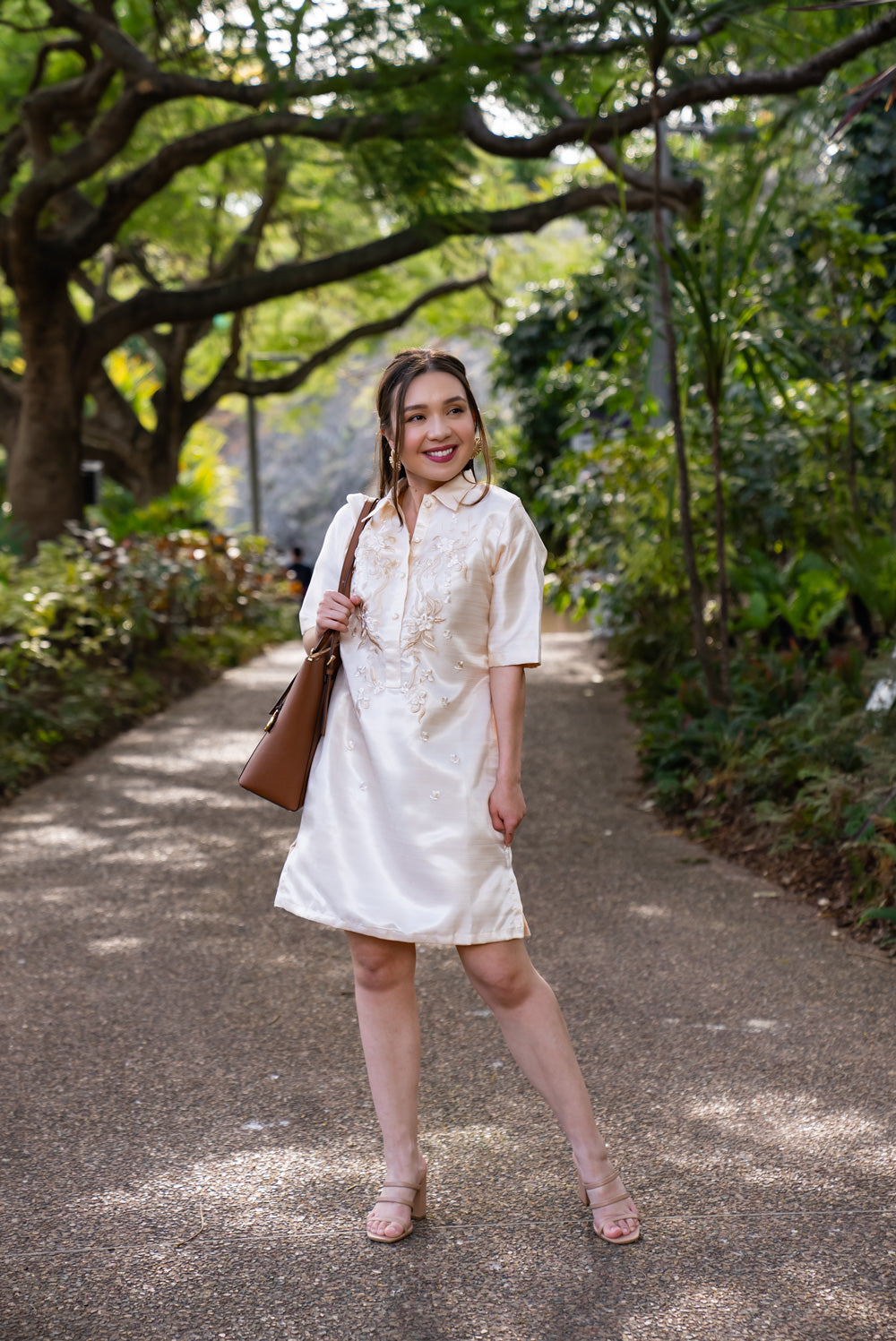 Model wearing the Amada Barong Dress in cream, a Filipiniana shift dress with hand embroidery, styled with nude heels and a tan handbag