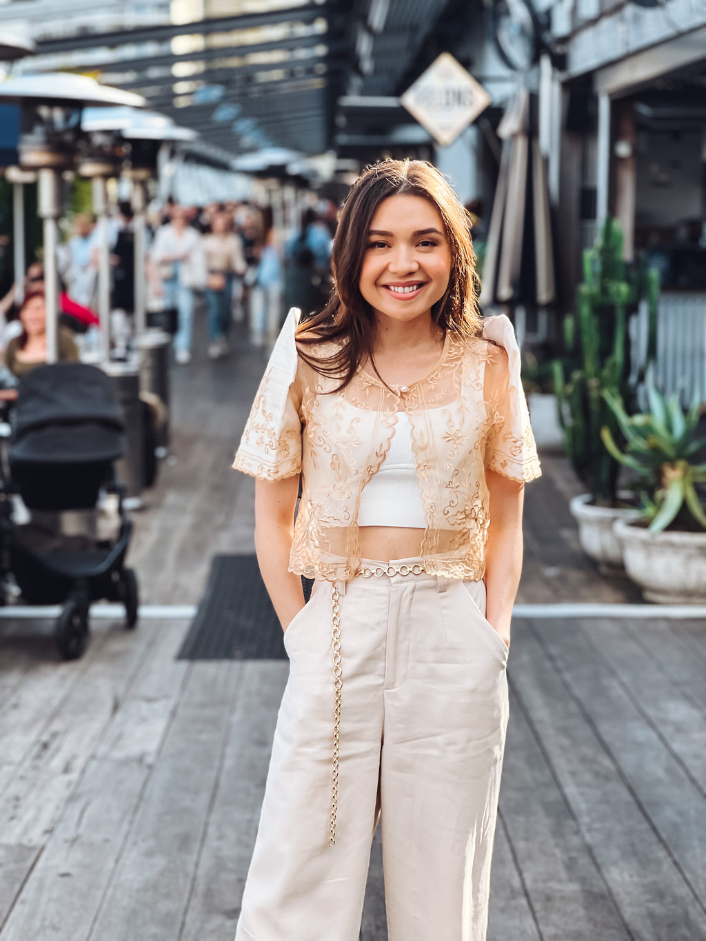 A young woman with brown hair smiles on a wooden outdoor walkway, wearing the Carmen Mestiza Bolero – Cream from Mestiza Filipina with wide-leg pants, surrounded by people and plants in the background.
