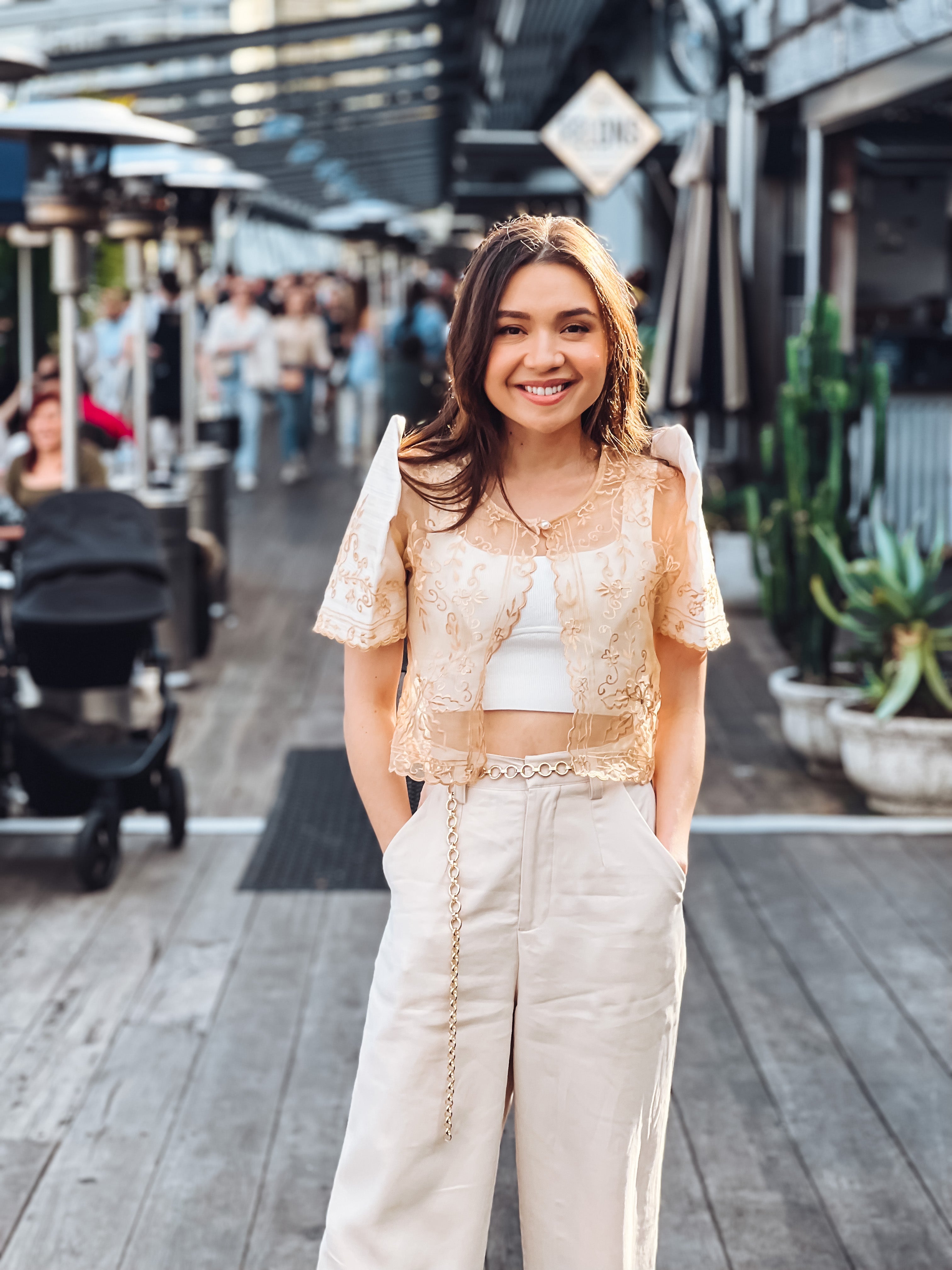 A young woman with brown hair smiles on a wooden outdoor walkway, wearing the Carmen Mestiza Bolero – Cream from Mestiza Filipina with wide-leg pants, surrounded by people and plants in the background.