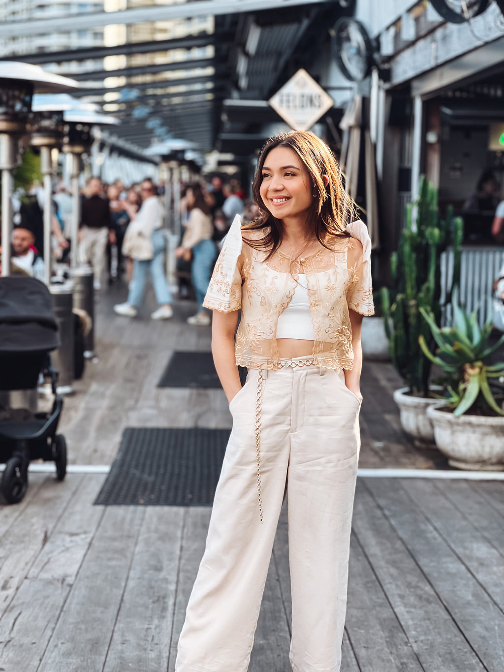 A young woman smiles on a wooden boardwalk, wearing the Mestiza Filipina Carmen Mestiza Bolero – Cream with hand embroidery and butterfly sleeves, surrounded by crowds, shops, and potted plants in the background.